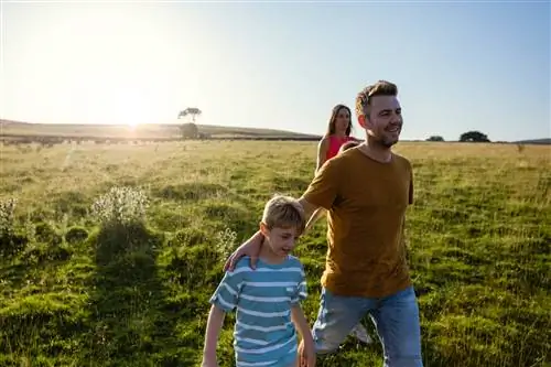 Папа гуляет с сыном по травяному полю, на заднем плане его жена.