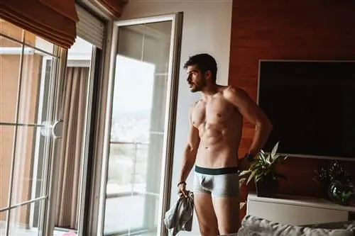 Мужчина в своей спальне носит только нижнее белье, штаны держит в руке.