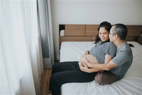 Мужчина сидит позади своей беременной жены на их кровати, обнимая ее.