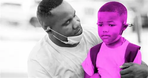 Отец в маске вокруг подбородка приседает и обнимает своего ребенка без маски.