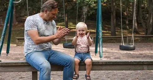 Папа и ребенок на скамейке на детской площадке хлопают в ладоши.