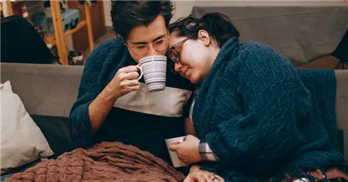 Супружеская пара с гриппом отсиживается дома, закутавшись в одеяла и пьет чай.