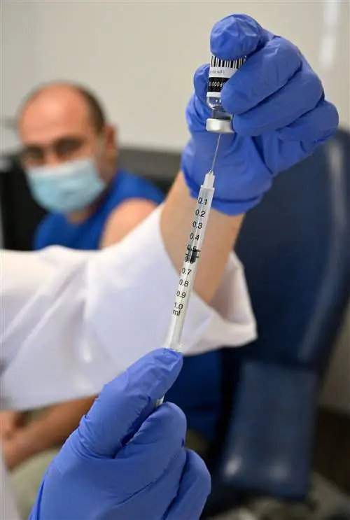 Медсестра готовит вакцину, а мужчина в маске ждет ее на заднем плане.