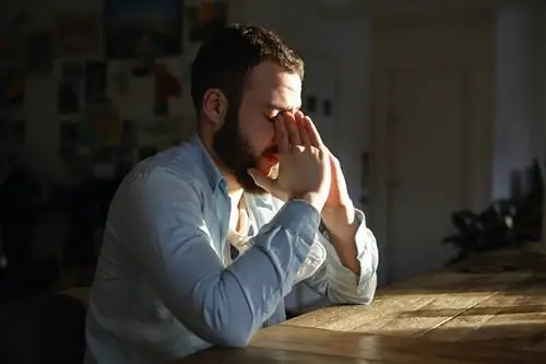Мужчина, сидящий за кухонным столом, выглядит встревоженным, закрыв лицо руками.