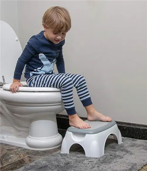 Маленький мальчик в пижаме сидит на унитазе с ногами на серо-белом табурете Nuby
