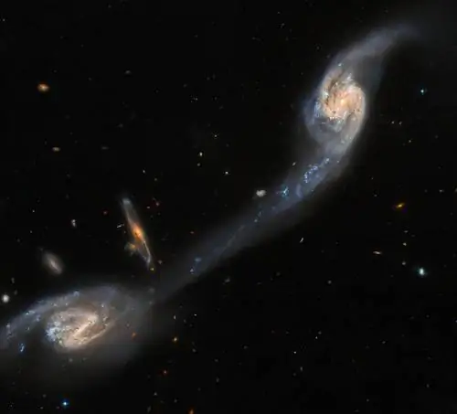 Две спиральные галактики видны почти лицом друг к другу; они представляют собой смесь бледно-голубого и желтого цвета, кро…